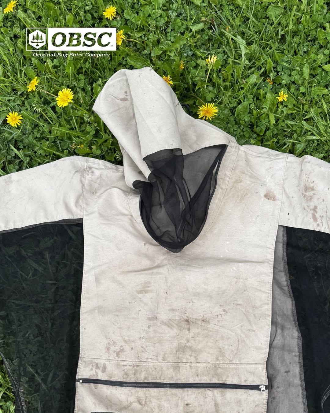 White Original Bug Shirt with black mesh panels and hood laid flat on green grass with dandelions